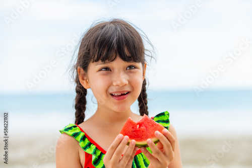 Little Asian child girl in swimsuit eating watermelon during playing with family on tropical beach in sunny day. Happy children kid enjoy and fun outdoor activity lifestyle on summer holiday vacation.
