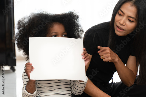 A black child is holding up a blank white piece of paper. sitting next to his mother in the living room of the house