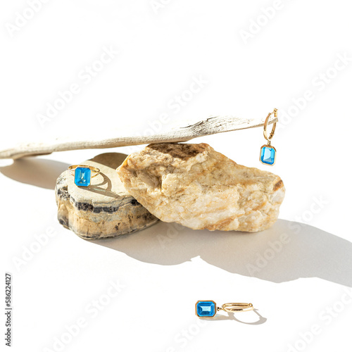 Elegant jewellery set of golden ring andearrings with blue topaz on white background with stones and wood. Minimal style composition with sunlit and shadow. Product still life concept. selective focus
