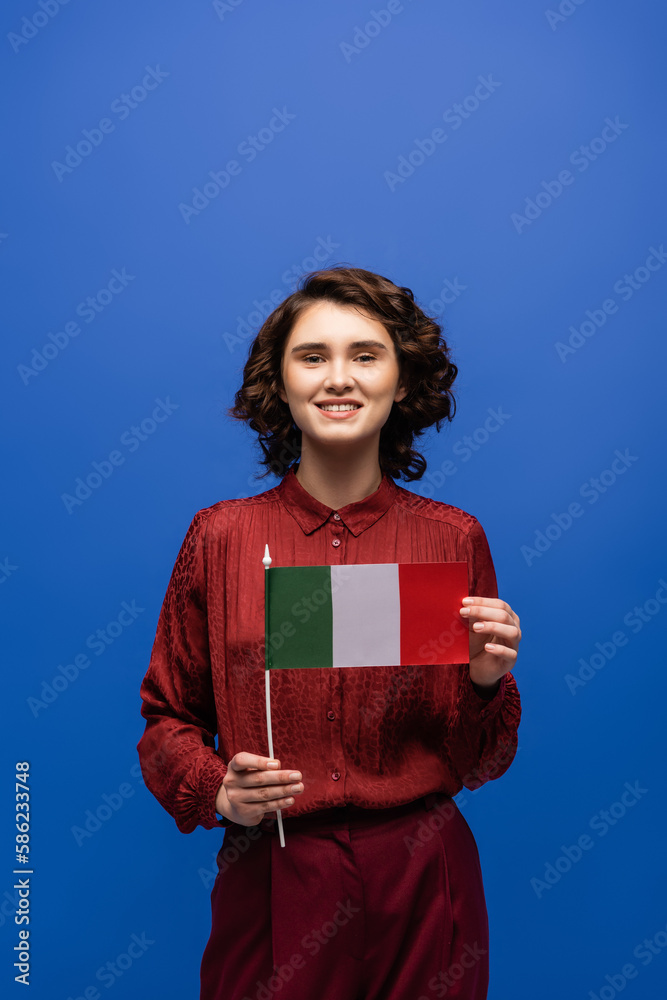 Obraz premium happy language teacher smiling while holding flag of Italy and looking at camera isolated on blue.