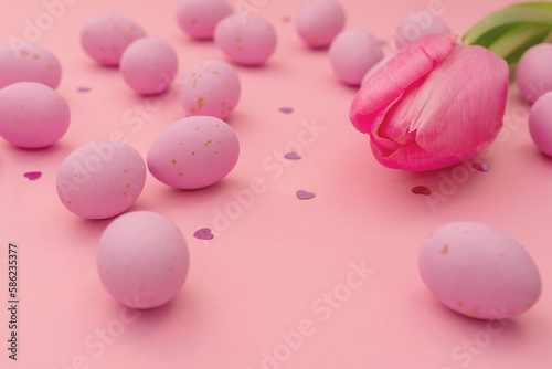 Easter greeting card. Close-up of bright pink chocolate delicious eggs scattered on pink background. Shimmering shining hearts and delicate tulip. Use in marketing materials, packaging. Happy holidays