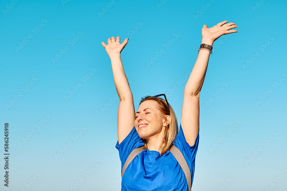 Blond woman tourist with closed eyes spreading arms up to sky enjoying freedom by the sea. Female traveler relaxing in serene nature. Mental health, wellness, adventure travel and healthy lifestyle.