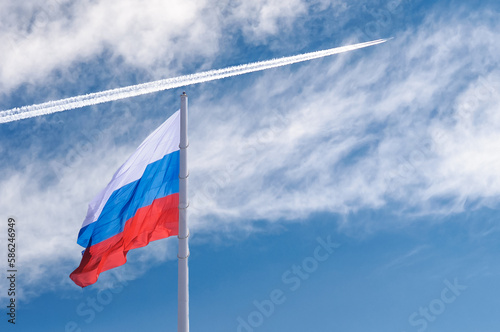 Flag of Russia against a blue cloudy sky with a white trail from the plane. State flag of the Russian Federation in the sun