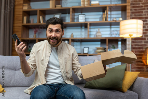 Portrait of happy online shopper at home, mature adult man smiling and looking at camera, holding product box and phone, happy about fast delivery from online store.