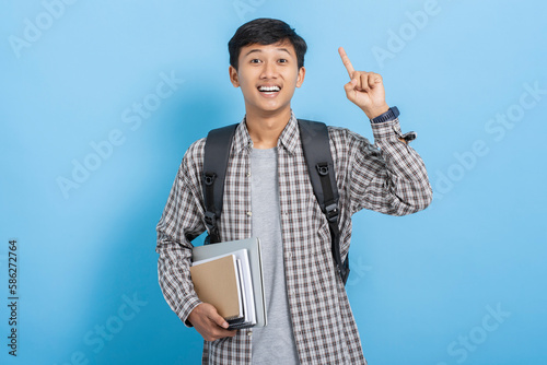 Young asian college student carrying books and backpack looking up thinking expression isolated over blue background