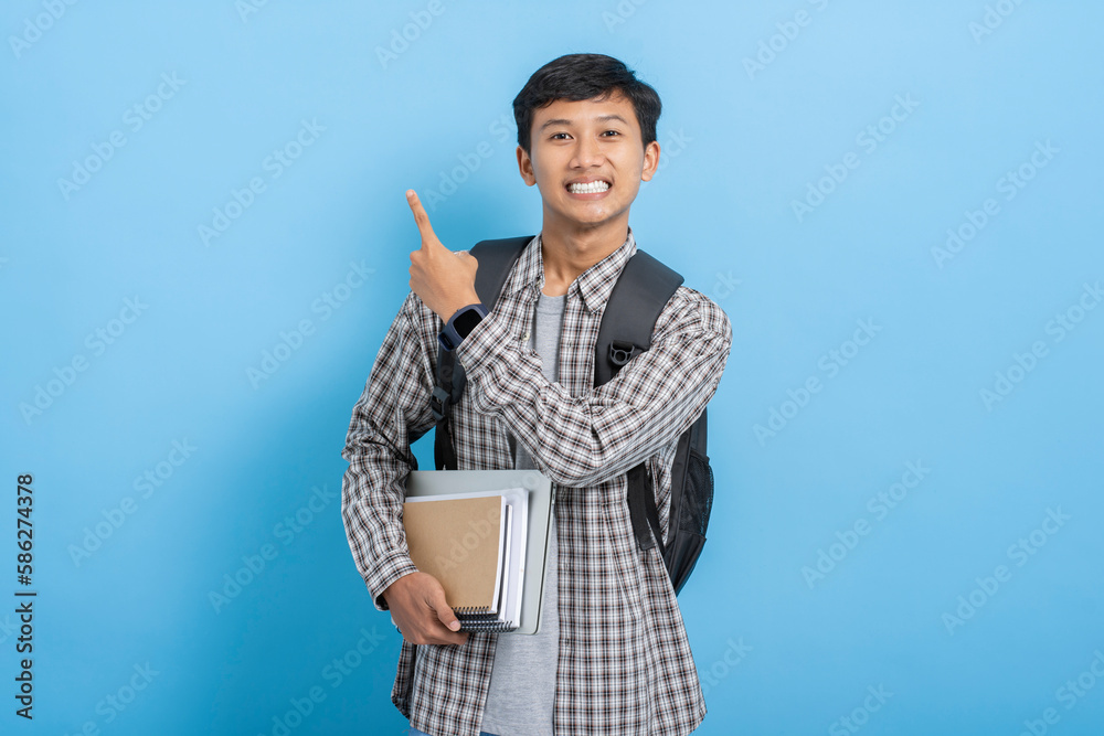 Young asian college student carrying books and backpack pointing with finger on blank copyspace isolated over blue background Young asian college student carrying books and backpack pointing with finger on blank copyspace isolated over blue background
