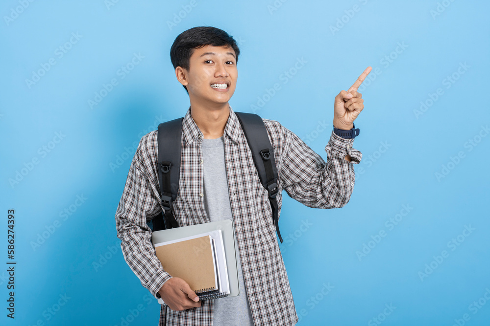 Young asian college student carrying books and backpack pointing with finger on blank copyspace isolated over blue background Young asian college student carrying books and backpack pointing with finger on blank copyspace isolated over blue background
