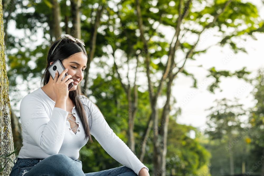 beautiful young latin college girl sitting in a green park talking on the cell phone