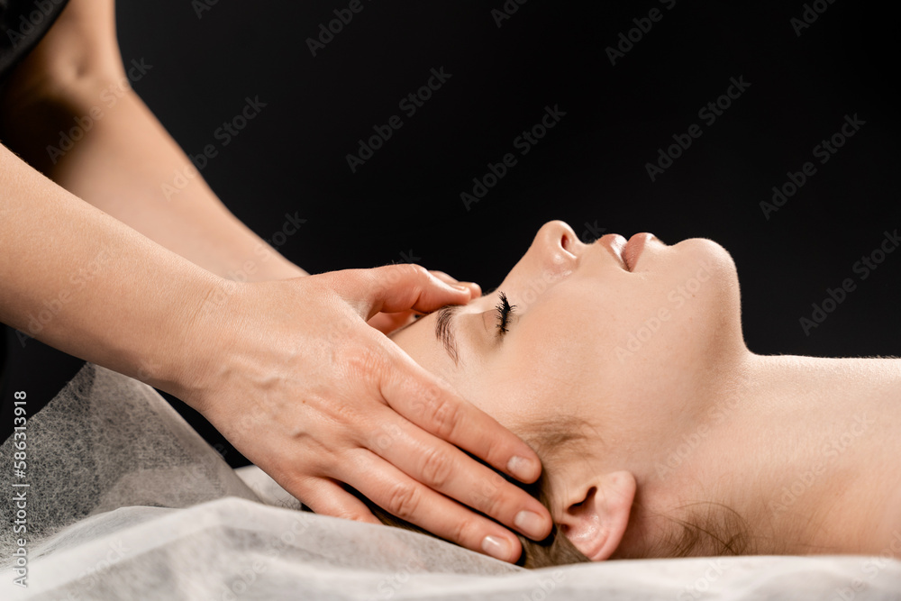 Girl with closed eyes is relaxing on face and neck massage in spa. Masseur is making facial massage for young woman in spa. Facebuilding and relaxing procedures.