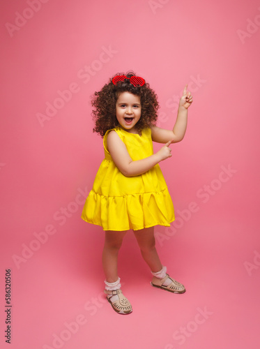 cute attractive joyful little girl in yellow dress pointing her index finger up.pink pastel color background