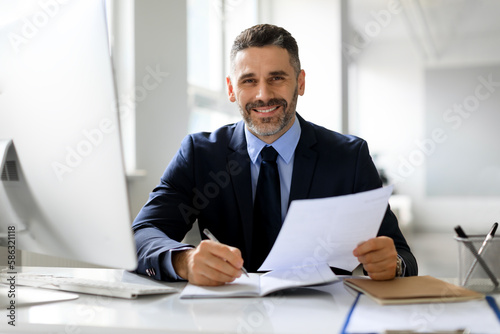 Happy male entrepreneur checking financial reports at desk in office and smiling at camera, enjoying company growth