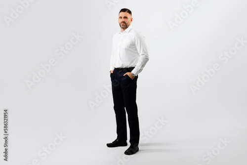 Full body length shot of middle aged male entrepreneur holding hands in pockets, posing over light grey background