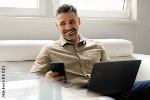 Happy middle aged male ceo holding smartphone and using laptop, sitting in office interior on couch, free space