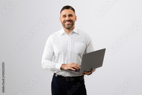 Portrait of cheerful middle aged businessman with laptop in hands smiling at camera, standing on light grey background
