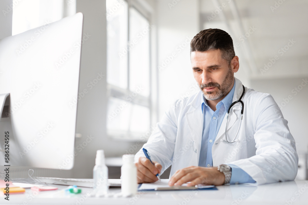Serious male doctor using computer and writing notes in medical journal sitting at workplace in clinic