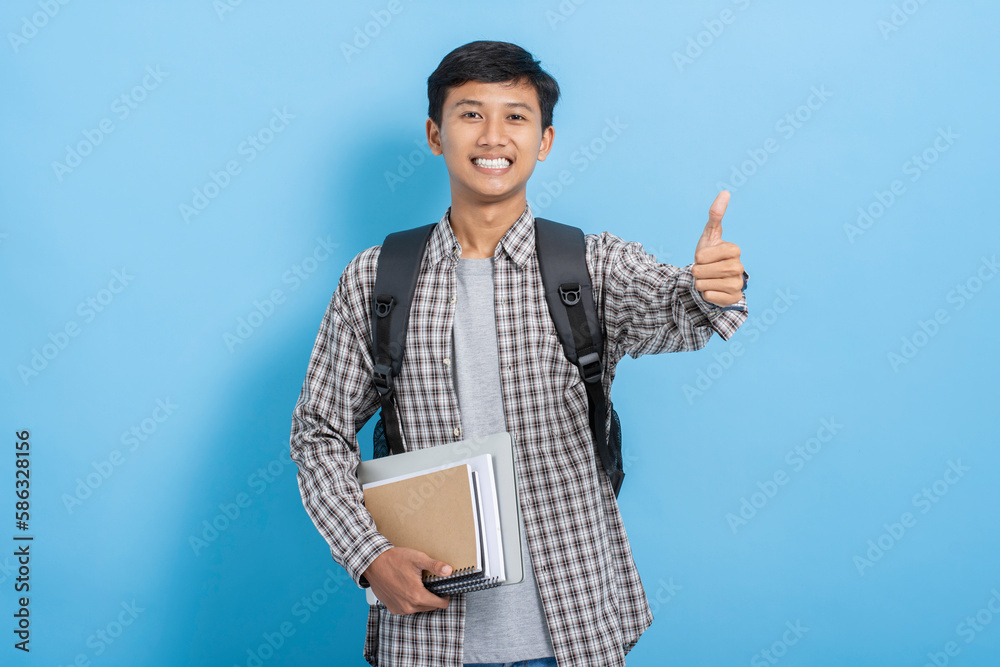 Young asian college student carrying books and backpack expressing positive doing thumbs up gesture isolated over blue background