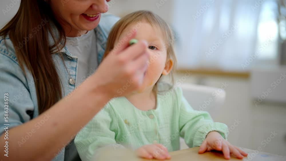 Mother feeds her baby toddler dairy food yogurt from plastic container on kitchen at home sitting at table. Mom holds her little kid with dirty face on knees. Raising child, kid's nutrition concept. Mother feeds her baby toddler dairy food yogurt from plastic container on kitchen at home sitting at table. Mom holds her little kid with dirty face on knees. Raising child, kid's nutrition concept.