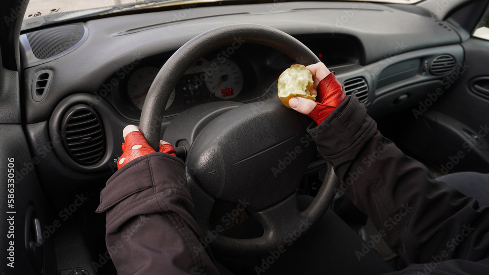  hands on the wheel of a car in red gloves, with an apple in one hand a person drives a car. Concept: Eating while driving a car   - obrazy, fototapety, plakaty 