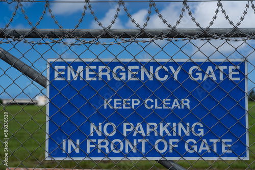 Airport Environment - Airport fence with a blue warning sign "EMERGENCY GATE", "KEEP CLEAR", "NO PARKING IN FRONT OF GATE" in front of blurred background 