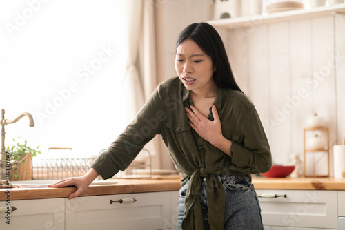 Sick chinese woman suffering from heartburn in kitchen, touching chest photo