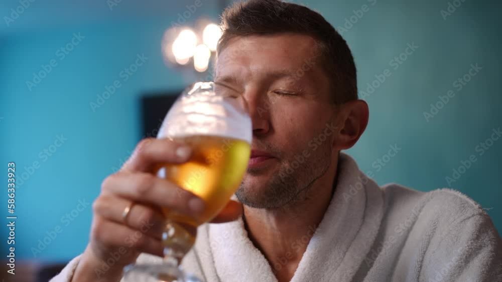 Front view portrait of confident handsome Caucasian man drinking refreshing beer indoors. Close-up satisfied male client in white bathrobe enjoying alcohol drink in SPA bar Front view portrait of confident handsome Caucasian man drinking refreshing beer indoors. Close-up satisfied male client in white bathrobe enjoying alcohol drink in SPA bar