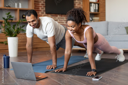 Smiling young black couple in sportswear watch video on laptop  doing push-ups on floor  workout together