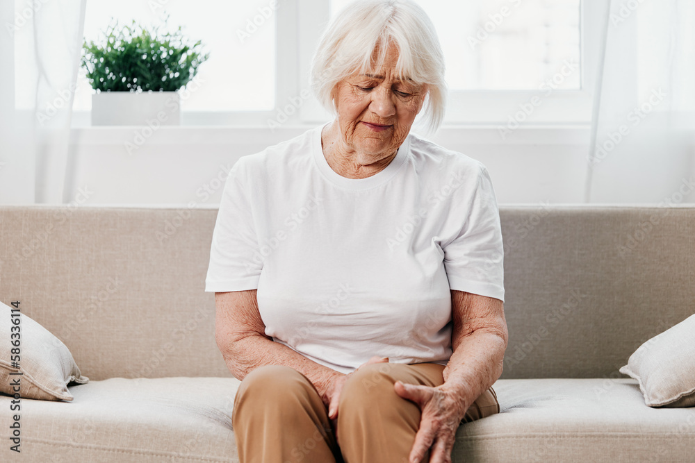 Elderly woman severe pain in her leg sitting on the couch, health problems in old age, poor quality of life. Grandmother with gray hair holds on to her sore knee, problems with joints and ligaments. Elderly woman severe pain in her leg sitting on the couch, health problems in old age, poor quality of life. Grandmother with gray hair holds on to her sore knee, problems with joints and ligaments.