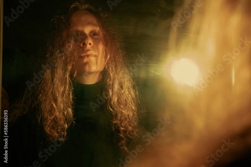 Portrait of young man with long blond curly hair under the light of lamp in the mirror. Solitude, bright loneliness, creativity, thoughtfulness © Annatamila