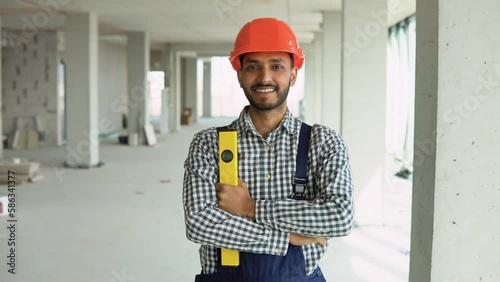 Portrait of Indian worker builder in uniform wearing a safety helmet with waterpas looking at the camera, standing on construction site of office center photo