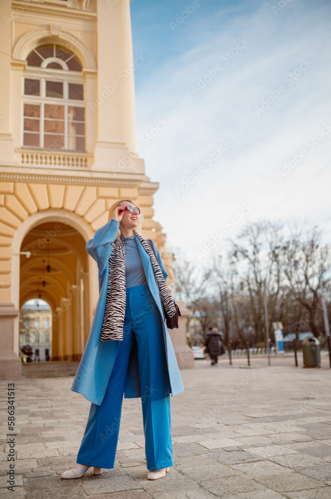 Happy smiling fashionable woman wearing trendy outfit with blue midi coat, turtleneck, wide leg trousers, zebra print scarf, posing in street of European city. Full-length outdoor portrait
