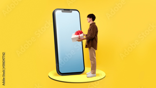 Man Holding Gift Box Near Phone Screen Over Yellow Background © Prostock-studio