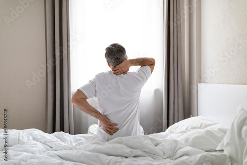 Unrecognizable grey-haired man sittting on bed, touching back and neck