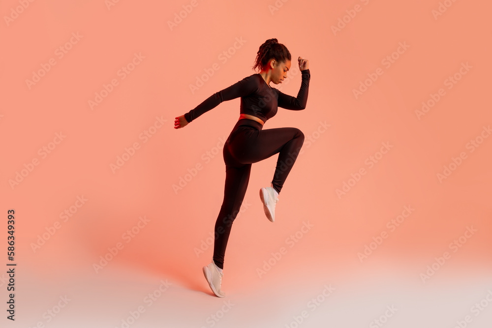 Athletic black lady jumping in mid air, training over peach neon studio background, full length, side view, copy space