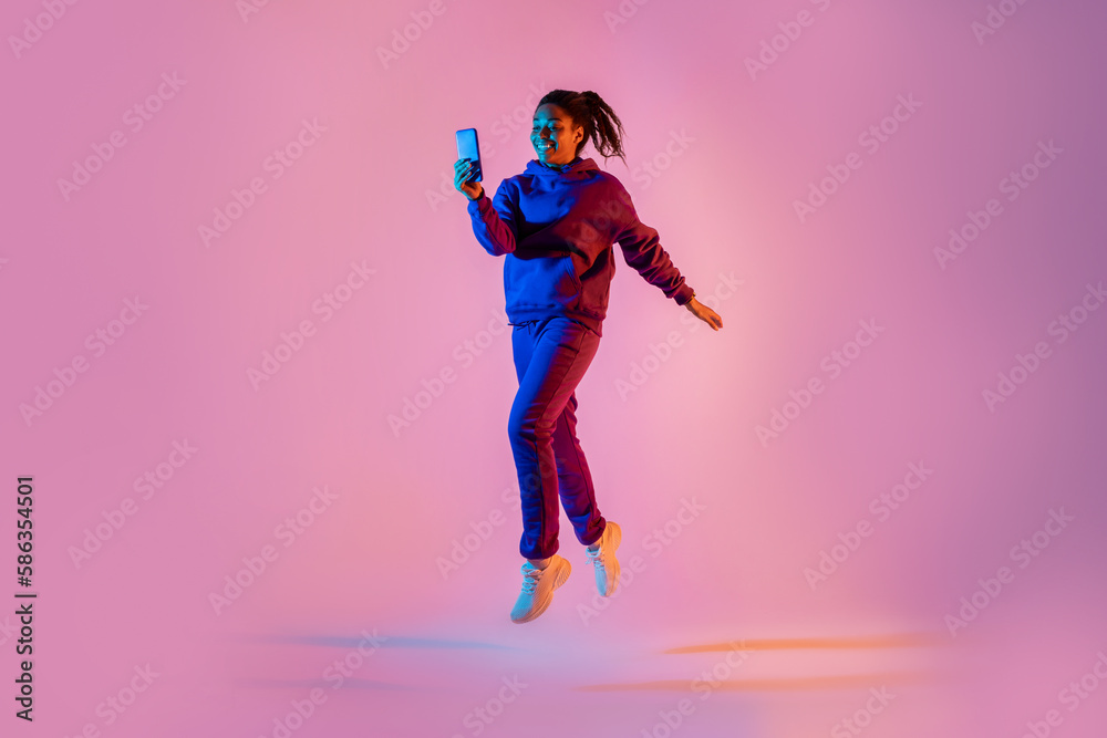 Full body length shot of young black lady in sportswear jumping and using cellphone over pink neon background