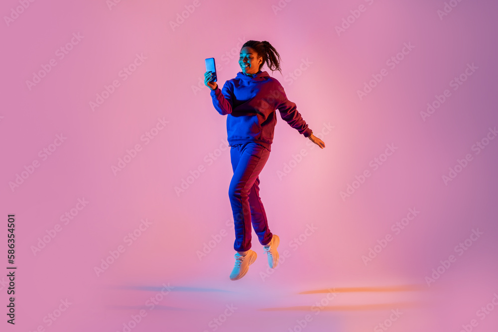 Full body length shot of young black lady in sportswear jumping and using cellphone over pink neon background Full body length shot of young black lady in sportswear jumping and using cellphone over pink neon background
