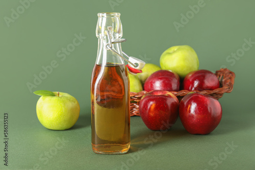 Glass bottle of fresh apple cider vinegar and basket with fruits on green background