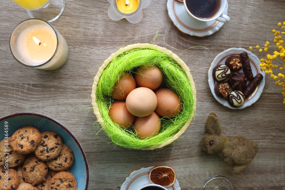 Easter eggs in the basket, bowl of cookies, chocolate pralines, Easter bunny figurine, cups of tea, glasses of juice, flowers and lit candles on the table. Top view. Easter eggs in the basket, bowl of cookies, chocolate pralines, Easter bunny figurine, cups of tea, glasses of juice, flowers and lit candles on the table. Top view.