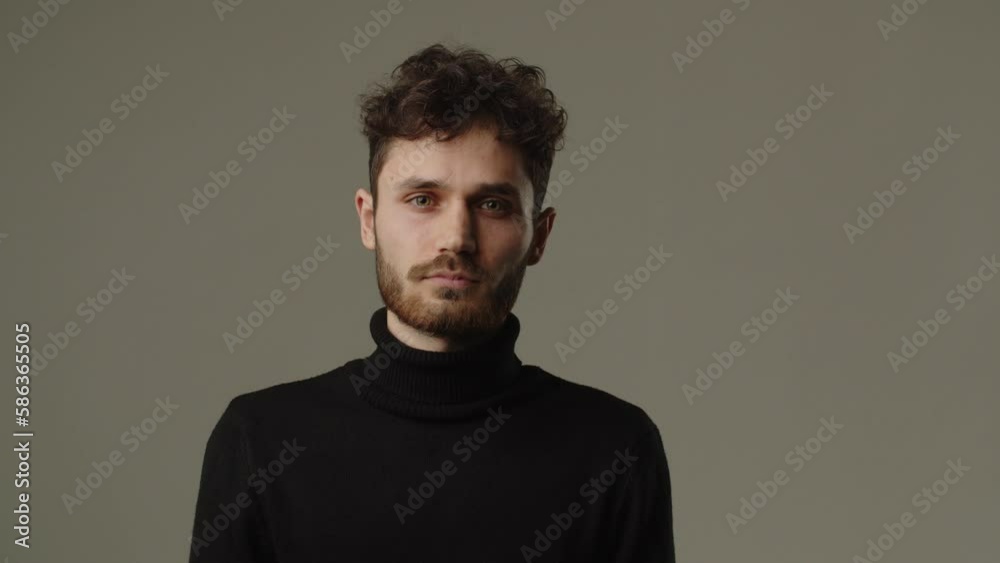 Front view of a man with curly hair and stubble on his face. Studio footage of serious guy looking at camera and blinking on grey background Front view of a man with curly hair and stubble on his face. Studio footage of serious guy looking at camera and blinking on grey background