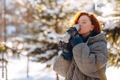 Portrait shot of a cute red-haired woman drinking tea from a thermos in the middle of a winter forest. A woman in a gray down jacket drinks hot tea from a thermos to keep warm after a long walk.