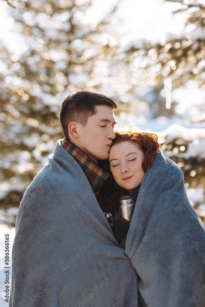 Young people in love stand in the middle of a sunny winter park wrapped in a blanket to keep warm. A young couple spends their day off walking in a snowy park. Young people in love stand in the middle of a sunny winter park wrapped in a blanket to keep warm. A young couple spends their day off walking in a snowy park.