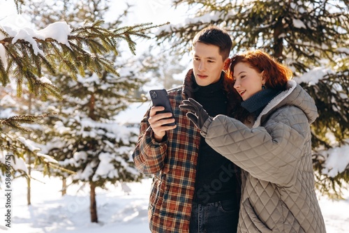 A young guy and a girl in winter clothes are looking at something on a smartphone. The guy and his girlfriend are looking at photos taken while walking through a snowy park.