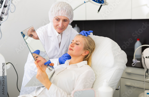 Professional female cosmetologist planning upcoming facial treatments for adult woman in modern aesthetic medicine office  patient looking at mirror