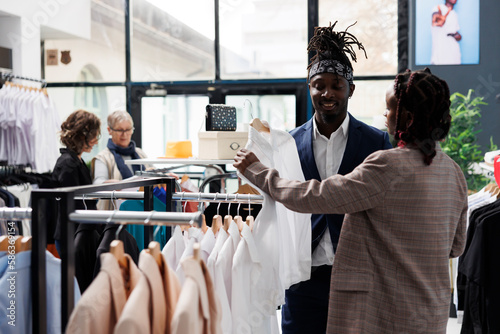 Showroom employee helping african american client with choosing white shirt during commerical activity. Elegant customer buying fashionable clothes in shopping centre. Fashion concept