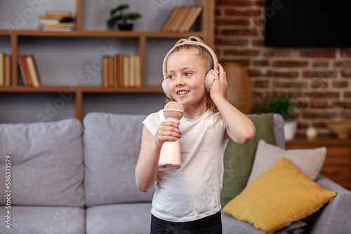 Child wearing headphones using microphone, singing song on sofa in room. Having fun at home.