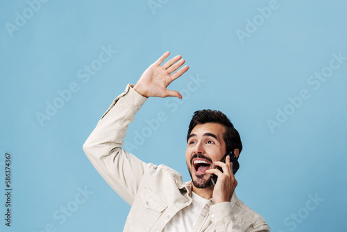 Portrait of a man brunette animation and joy talking on the phone hand and fist up from victory joy, on a blue background in a white T-shirt and jeans, copy space