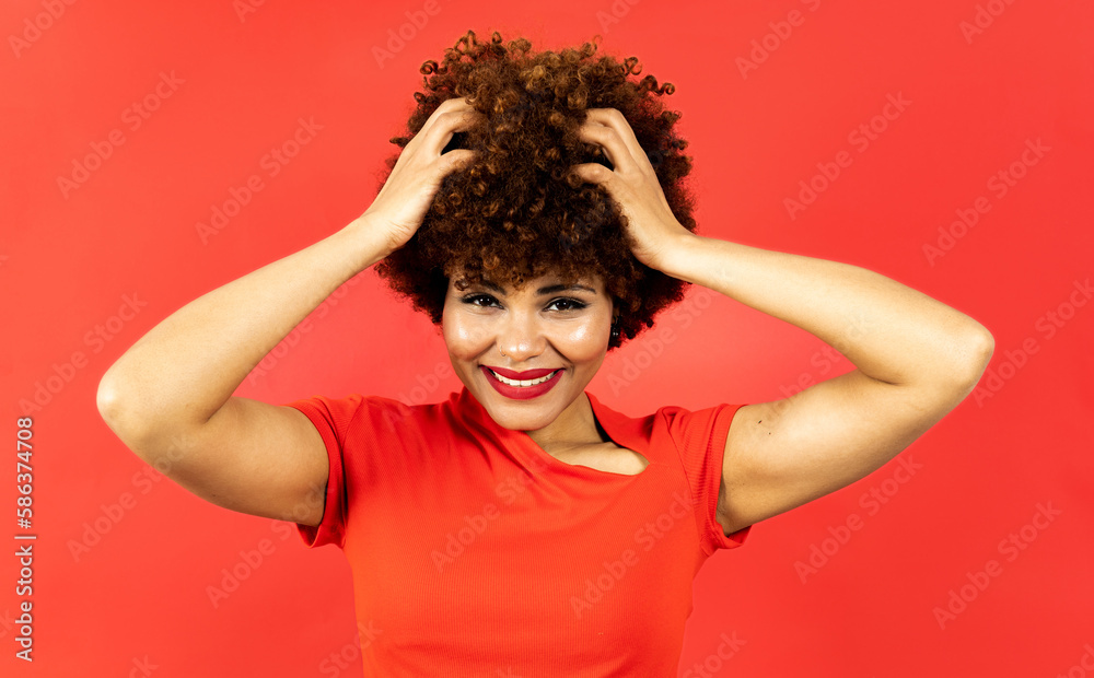 A dark-skinned woman with afro hair poses on a red studio background.The girl poses touching her hair with the expression of not being able to comb it. Concept of frizzy hair, rebellious hair. A dark-skinned woman with afro hair poses on a red studio background.The girl poses touching her hair with the expression of not being able to comb it. Concept of frizzy hair, rebellious hair.