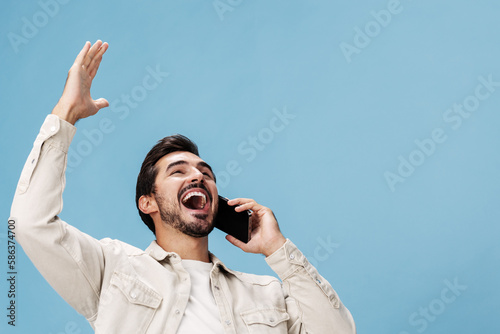 Portrait of a man brunette animation and joy talking on the phone hand and fist up from victory joy, on a blue background in a white T-shirt and jeans, copy space