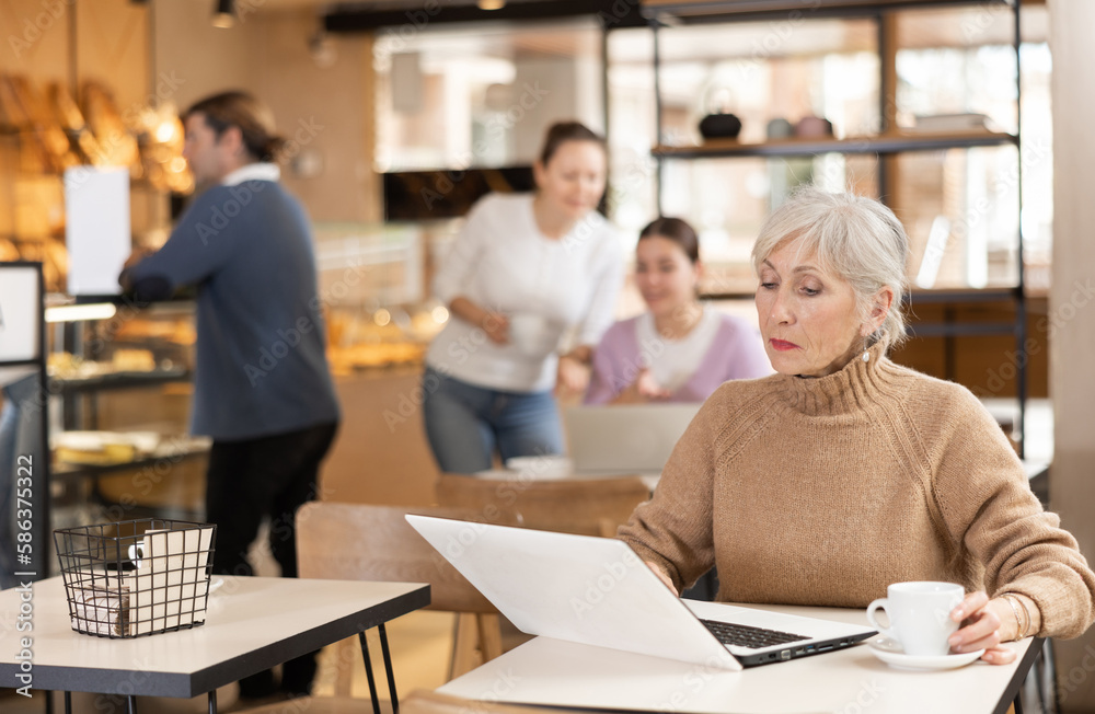 Positive modern aged female freelancer sitting at table in cafe with cup of coffee and laptop, browsing websites on internet
