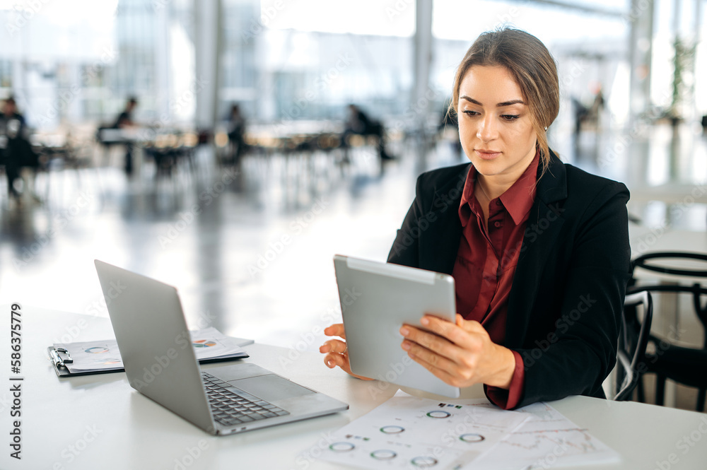 Focused elegant successful caucasian woman, entrepreneur, top manager, employee of marketing department, sits at a desk in the office, focused looks at a tablet screen, working on a project Focused elegant successful caucasian woman, entrepreneur, top manager, employee of marketing department, sits at a desk in the office, focused looks at a tablet screen, working on a project