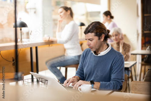 Positive European middle-aged man drinking and enjoying coffee while working on laptop in cafe. Freelance and remote work